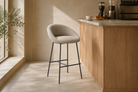 Dune beige curved-shell bar stool at a boutique café counter with structured woven upholstery and matte black H-bar footrest frame.