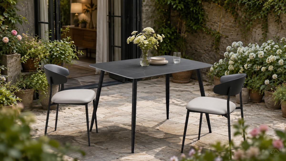 GEYA T-107 sintered stone dining table styled in a green courtyard dining setting with two chairs, flowering plants, Armani Grey top and matte black splayed legs.