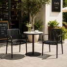 Matte black slat-back stackable polypropylene armchairs with integrated armrests in an outdoor café patio with a round bistro table and high-contrast natural daylight.