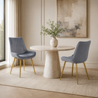 Grey-blue velvet diamond-quilted high-back dining chairs with gold tapered metal legs around a round stone dining table in a residential boutique dining room.